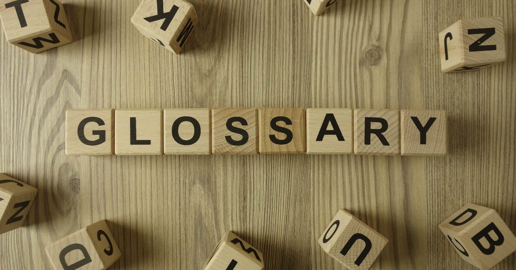 Image shows a wooden surface with wooden letters on it. The word Glossary is spelled out and the rest of the letters are just scattered randomly