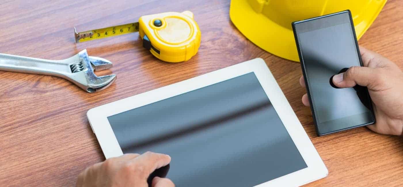 Photograph of a wooden surface with a spanner, tape measure and yellow hard hat at the back of the table. At the front of the table is a tablet with a hand reaching out to it and the other hand holding a cellphone