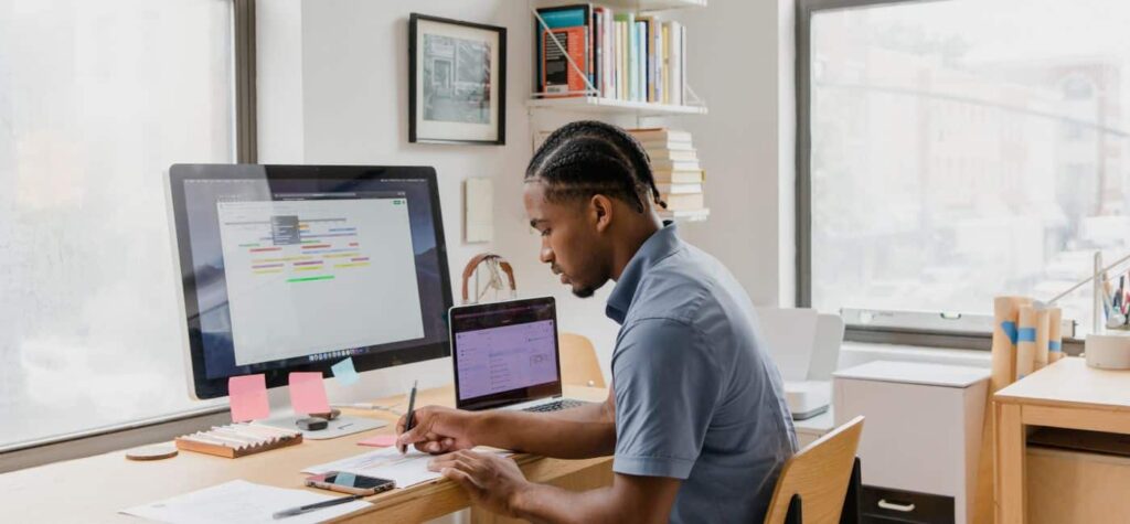 Black man in a light blue shirt sitting at a desk right him. In front of him is a computer monitor with some information on and next to that is a small laptop also with information on.